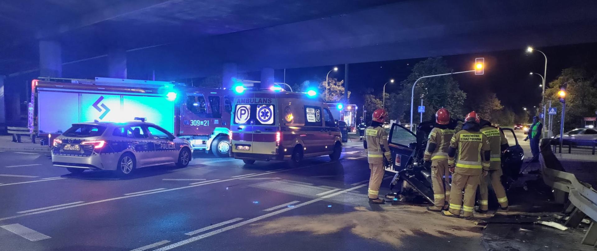 Na zdjęciu widoczni są pracujący strażacy oraz uszkodzony samochód osobowy. Widoczny jest samochód straży pożarnej, radiowóz policji oraz karetka pogotowia.