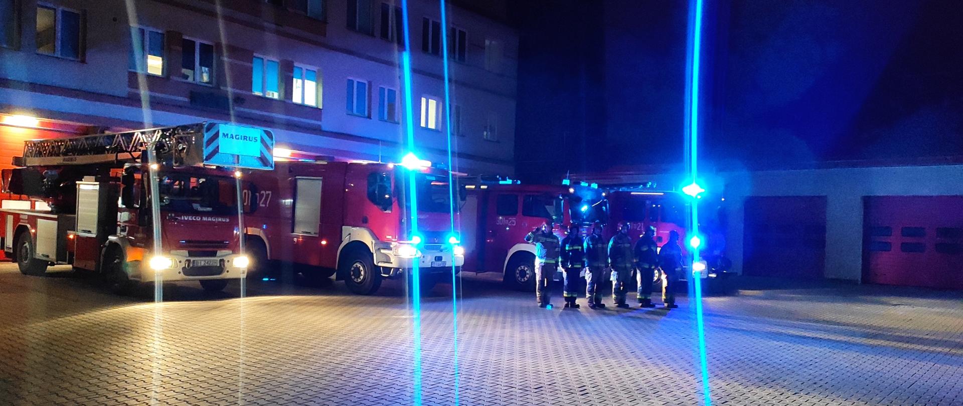 Widoczni strażacy na tle pojazdów gaśniczych i garaży, oddają honor
