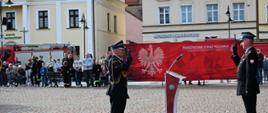 Na pierwszym planie, na wybrukowanym placu, stoi dwóch oficerów Państwowej Straży Pożarnej w ciemnych mundurach galowych, zwróconych do siebie i jednocześnie salutujących. Pomiędzy nimi znajduje się nowoczesna, czerwona mównica z mikrofonem. W tle widoczna jest duża czerwona ścianka promocyjna z białym orłem oraz napisem „Państwowa Straż Pożarna”. Za nią oraz po lewej stronie placu zgromadzeni są mieszkańcy obserwujący ceremonię, a w oddali widać zabytkowe kamienice oraz zaparkowany czerwony wóz strażacki.