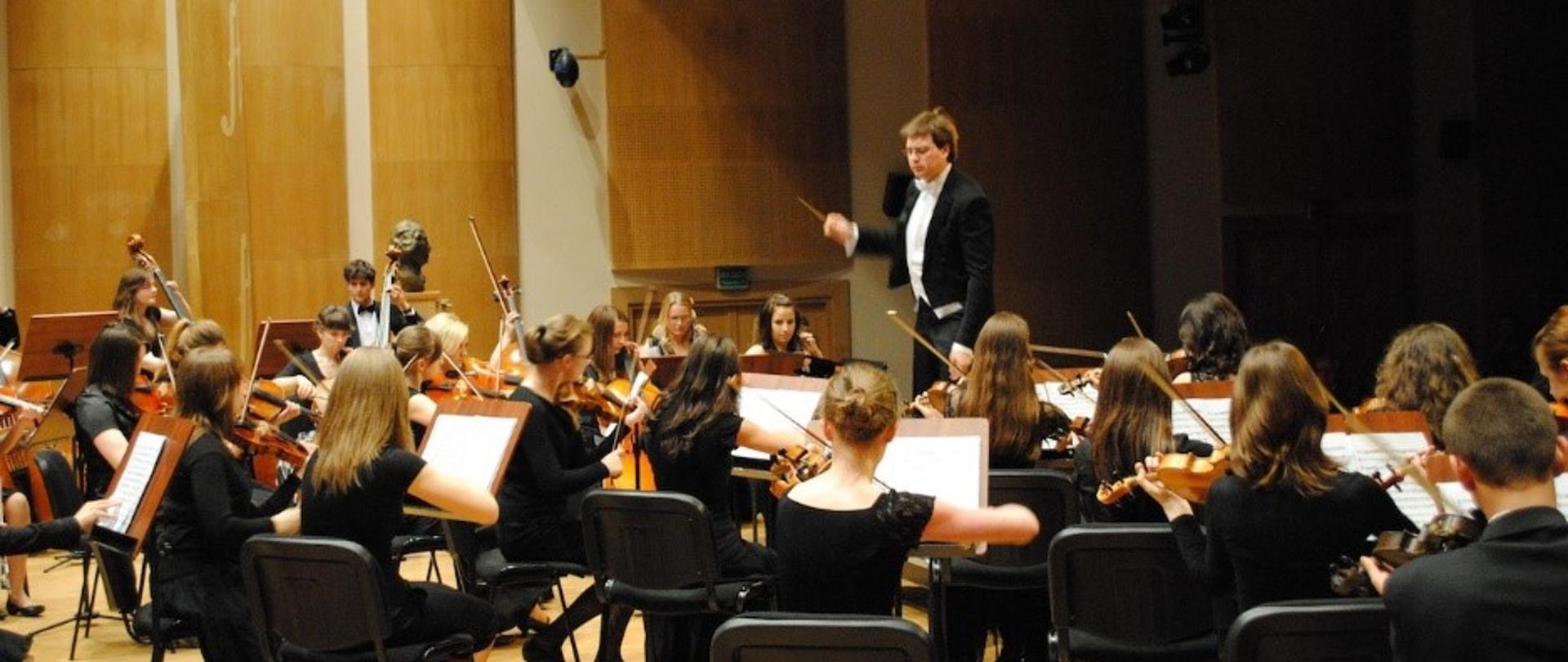 Grupa uczniów siedzących przy pulpitach, grających na instrumentach smyczkowych w czarnych strojach koncertowych. Przed nimi dyrygent z batutą ubrany w koncertowy frak.
