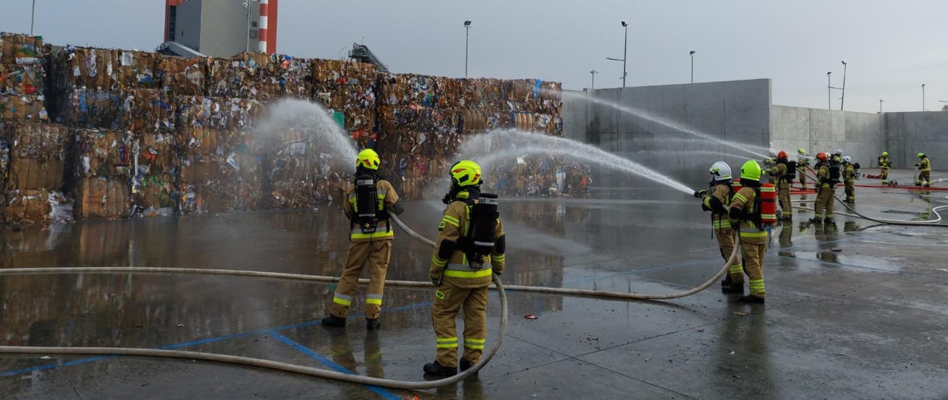 Na zdjęciu widać grupę strażaków w pełnym umundurowaniu bojowym, hełmach i aparatach oddechowych podczas ćwiczeń w składzie makulatury. Kilku strażaków trzyma węże i kieruje silne strumienie wody na wysokie bele sprasowanej makulatury ułożone w stos. Podłoże jest mokre, a w tle widać betonowe ściany oraz fragment zakładu przemysłowego z kominami.