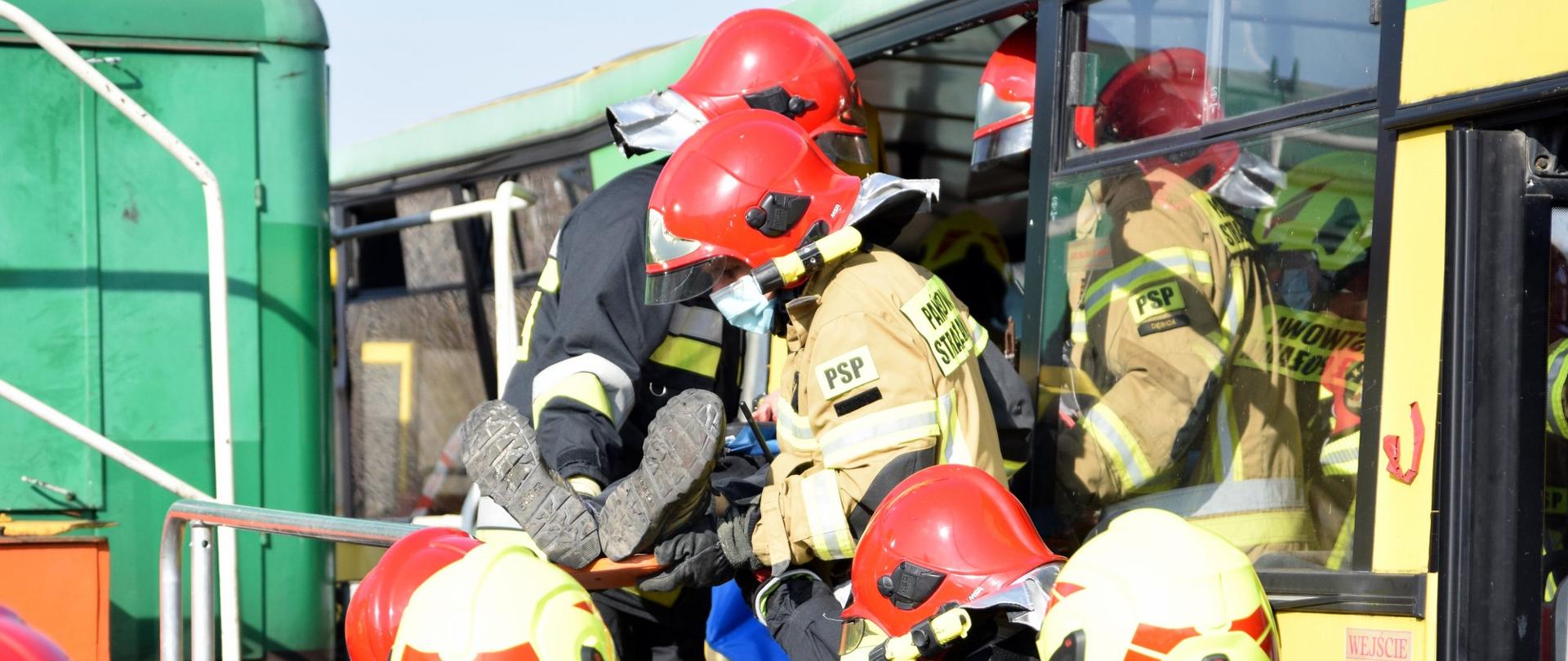 Strażacy ewakuują osobę poszkodowaną z uszkodzonego autokaru. Poszkodowany ułożony jest na desce ortopedycznej. Ewakuacja prowadzona jest przez okno autokaru. 