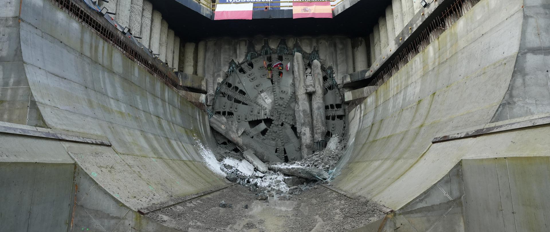 Zdjęcie przedstawia ogromną tarczę maszyny drążącej tunel (TBM) przebijającą się przez betonową ścianę w głębokiej niecce. Wokół tarczy leżą odłamki betonu, a nad nią widoczne są dwa banery z nazwami firm „Mostostal” i „Acciona”.