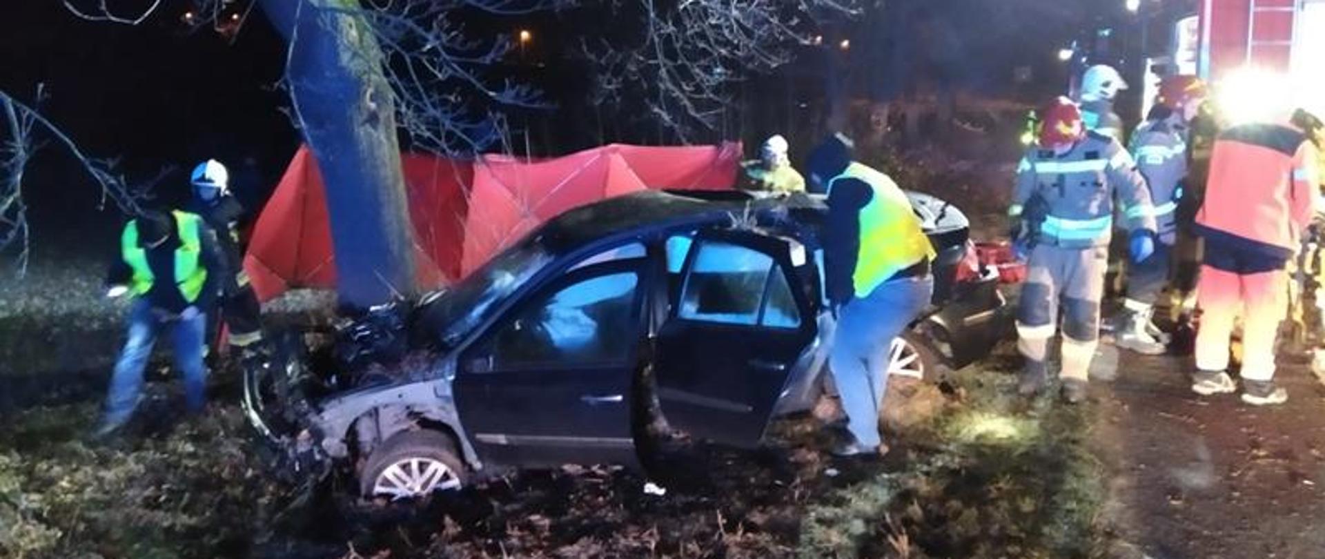 Zdjęcie przedstawia rozbity samochód osobowy stojący w rowie przy drzewie. Widok lewego boku rozbitego samochodu. Przy samochodzie funkcjonariusze policji wykonują czynności wyjaśniające, obok strażacy i ratownicy medyczni.