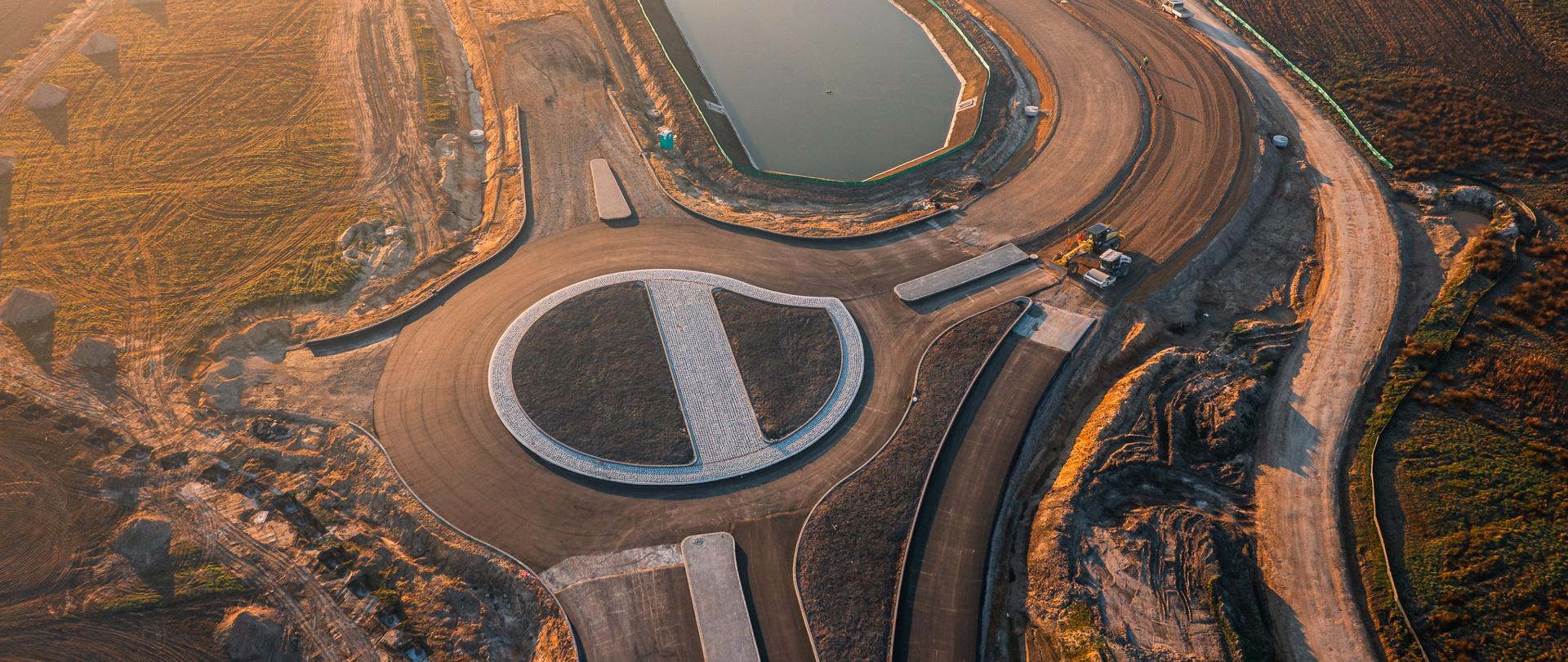 Zdjęcie przedstawia plac budowy z lotu ptaka. W centralnej części znajduje się nowo wybudowane rondo o kolistym kształcie, z brukowanymi elementami w środku. Wokół niego rozciągają się świeżo położone drogi, które są jeszcze w trakcie budowy. W tle widoczny jest duży, prostokątny zbiornik wodny, otoczony siecią dróg. Na placu budowy można dostrzec maszyny oraz pracowników, a cały obszar otoczony jest terenami zielonymi i polami. Zdjęcie zostało wykonane o zachodzie słońca, co nadaje mu ciepłe, złote światło.