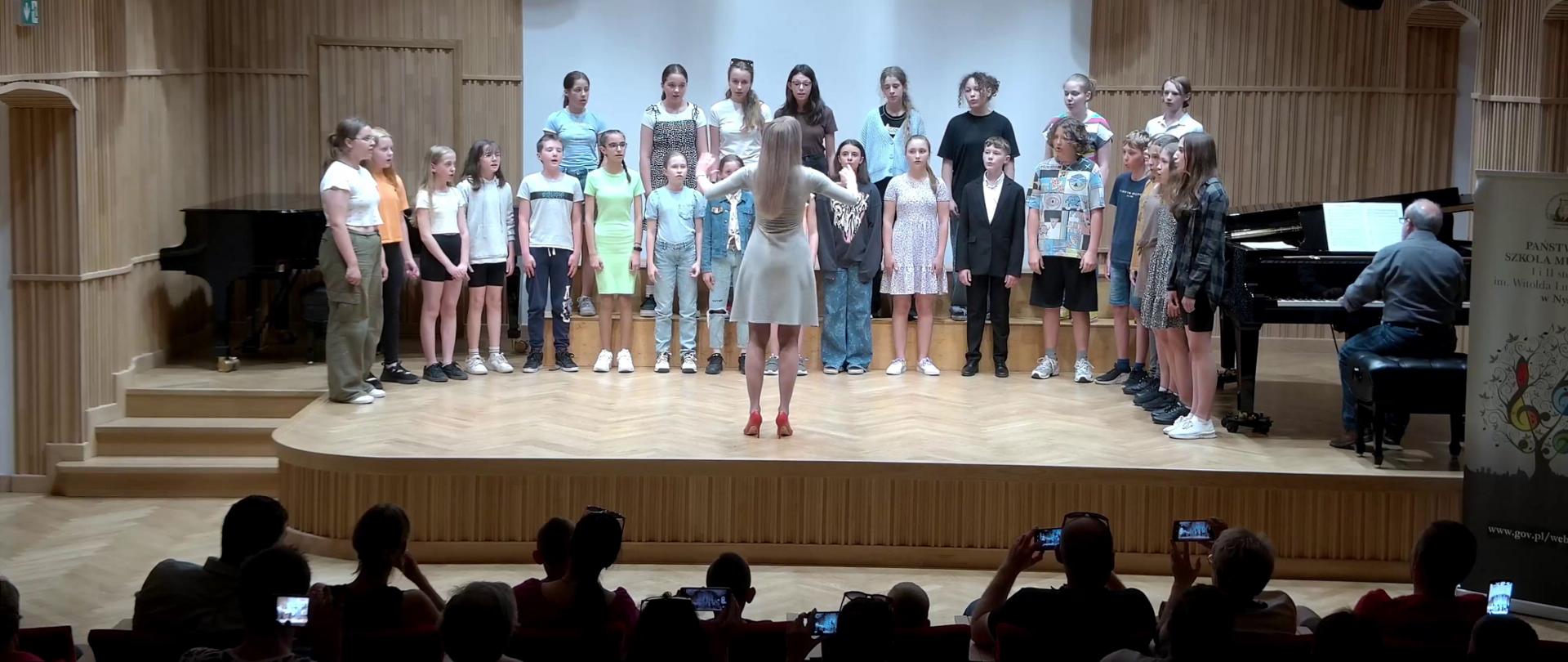 Zdjęcie. Na estradzie sali koncertowej występuje chór dziecięcy.. Z prawej strony widać również pianistę akompaniującego chórowi. Zespołem dyryguje pani dyrygent. Na dole zdjęcia widać publiczność, niektóre osoby nagrywają występ telefonami komórkowymi.