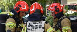 Trzech strażaków Państwowej Straży Pożarnej w piaszczysto-beżowych mundurach, aparatach oddechowych i czerwonych hełmach stoi blisko siebie i rozmawia. Strażak stojący tyłem w centrum uwagi nosi kamizelkę z czarnym napisem „KIERUJĄCY DZIAŁANIEM RATOWNICZYM”. W tle widać zielone drzewa oraz fragment czerwonego wozu strażackiego.
