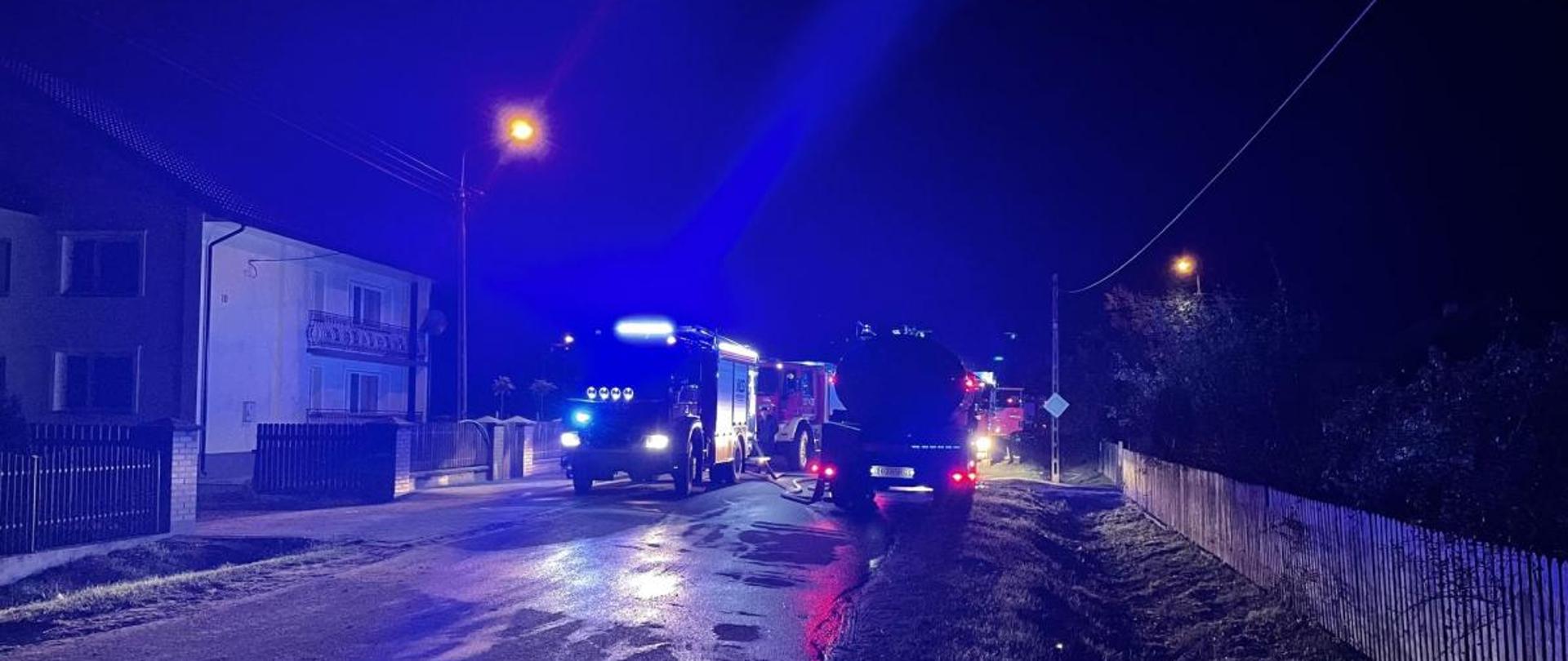 Zdjęcie przedstawia samochody bojowe straży pożarnej stojące na drodze z włączonymi światłami alarmowymi. Zdjęcie wykonano w zmroku.