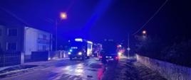 Zdjęcie przedstawia samochody bojowe straży pożarnej stojące na drodze z włączonymi światłami alarmowymi. Zdjęcie wykonano w zmroku.