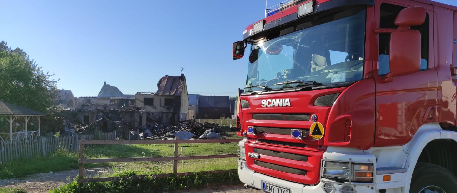 Zdjęcie przedstawia zniszczone w wyniku pożaru zabudowania mieszkalne i gospodarcze w miejscowości Nowa Biała. Po prawej stronie stoi samochód strażacki z JRG Myślenice