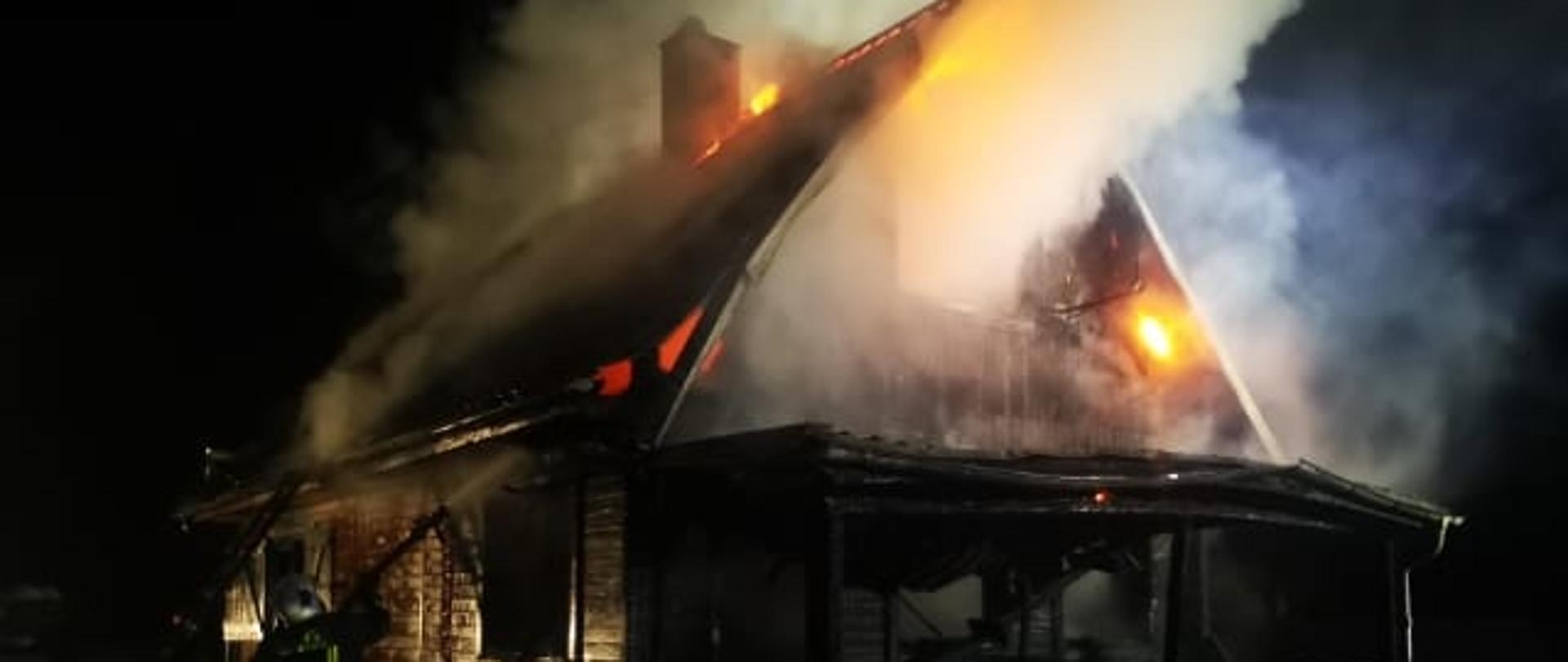 Pora nocna, bryła spalonego drewnianego budynku mieszkalnego, z okien wydostające się kłęby dymu, jak i delikatne zarzewia ognia. 