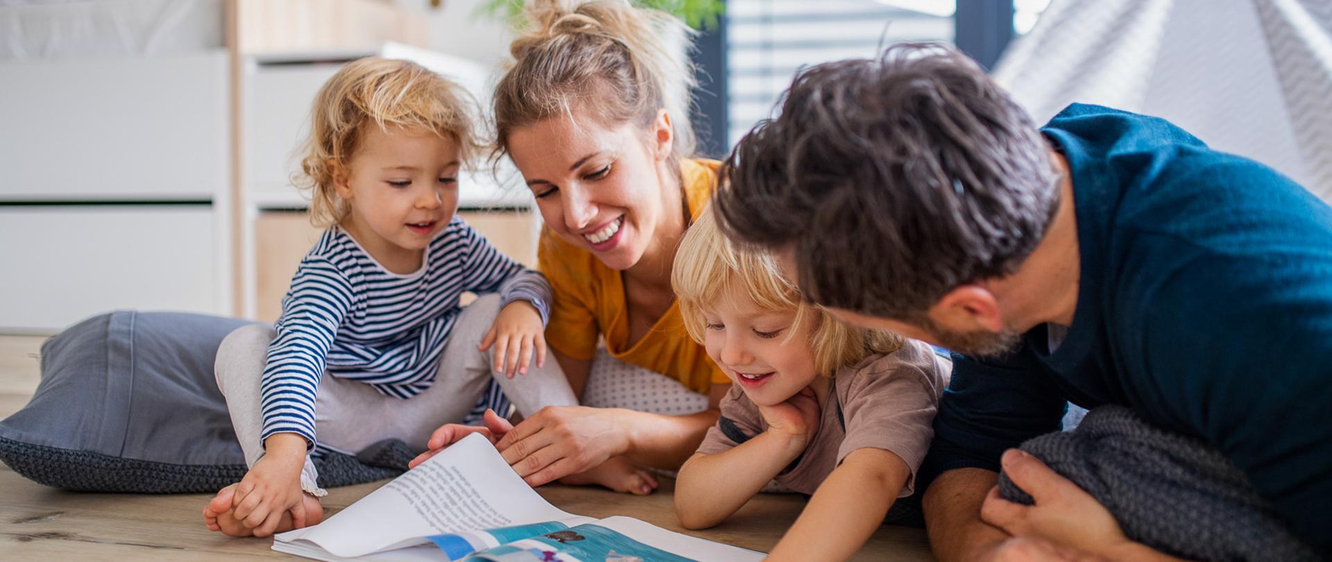 Front view of young family with two small children indoors in bedroom reading a book.