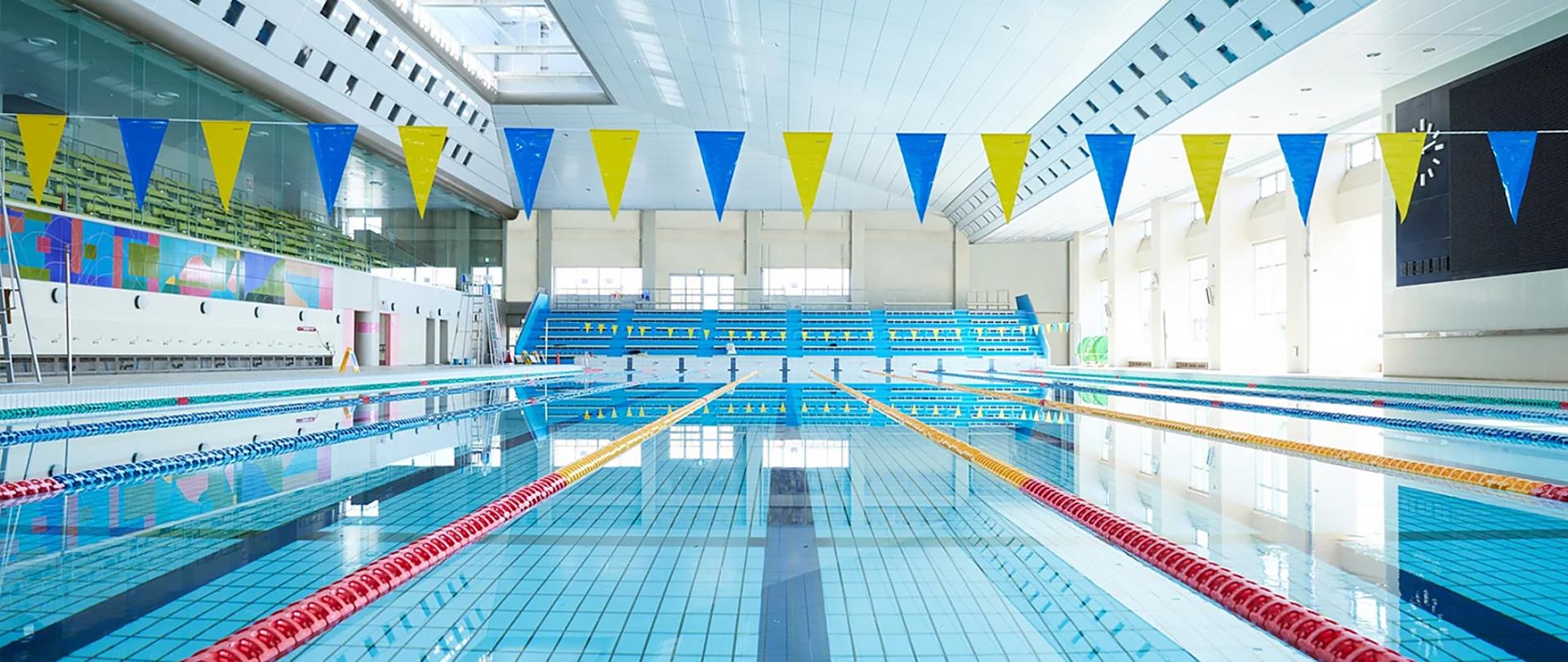 Na zdjęciu wnętrze nowoczesnego krytego basenu pływackiego z niebieską wodą, torami oddzielonymi kolorowymi bojkami oraz żółto-niebieskimi proporczykami zawieszonymi nad wodą. W tle widoczne są trybuny i duże okna wpuszczające naturalne światło.