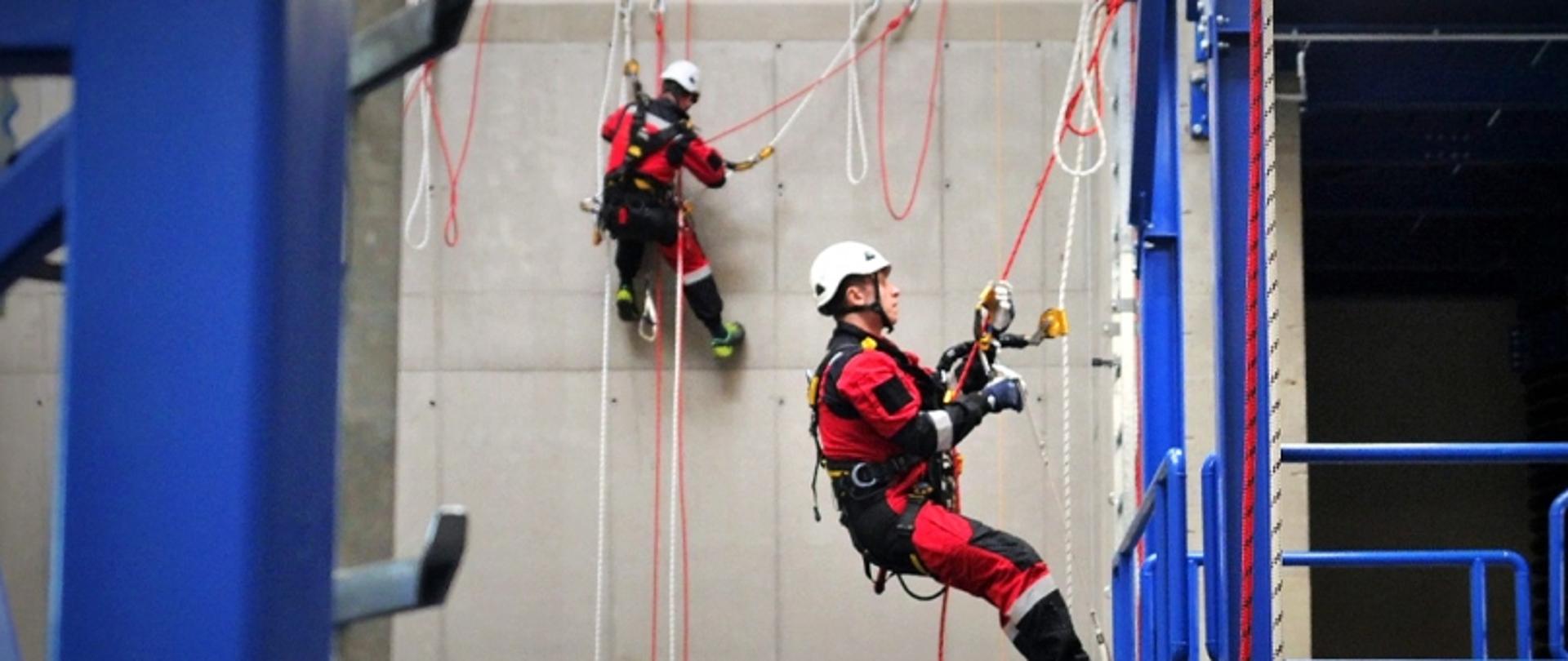 Na zdjęciu widać dwóch ratowników w czasie treningu wysokościowego. Ubrani są w czerwone kombinezony, na głowi maja białe kaski. Wiszą na linach.