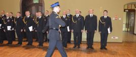 Dzień Strażaka w Rawiczu. Sala widowiskowa w domu kultury. Dowódca uroczystości maszeruje na tle pododdziału strażaków.
