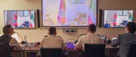 Czterech oficerów Krajowego Centrum Koordynacji Ratownictwa i Ochrony Ludności siedzących za stołem konferencyjnym podczas wideokonferencji. W tle monitory na których widoczni są strażacy z Kuwejtu. 