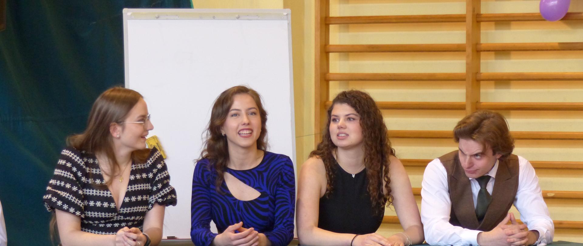 Na zdjęciu znajduje się grupa absolwentów biorących udział w części artystycznej uroczystości - Wielka Gra.
Absolwenci siedzą za stołem ubranym w zielony obrus. Od lewej strony: Anna Lenart, Emilia Wojtyczko, Małgorzata Sularz i Tadeusz Kmiecik