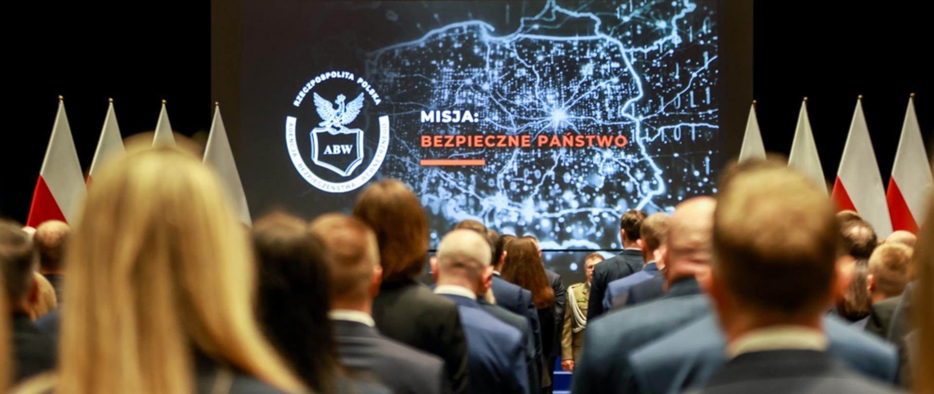 Zdjęcie przedstawia promocję funkcjonariuszek i funkcjonariuszy Agencji Bezpieczeństwa Wewnętrznego na pierwszy stopień oficerski. Promowani stoją tyłem. W tle na ekranie wyświetlone kontury Polski z podświetlonymi miastami i drogami i napisem MISJA: BEZPIECZNE PAŃSTWO. Po lewej stronie wyświetlono logo ABW (orzeł trzymający kontur Polski z napisem ABW). U góry zdjęcia widać czarne reflektory. Po prawej i lewej stronie ekranu widać stojące biało-czerwone flagi Polski - po cztery z każdej strony.