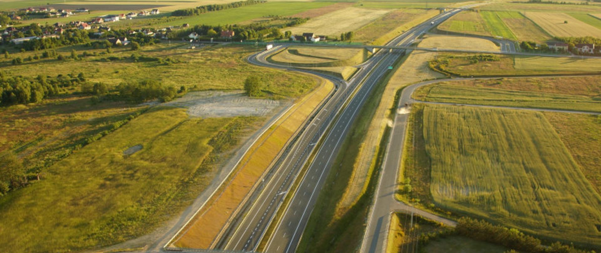 Zdjęcie lotnicze drogi ekspresowej S6. Widoczne dwie jezdnie drogi, węzeł z wiaduktami oraz drogami dojazdowymi. W tle widoczne zabudowania oraz farma wiatrowa.