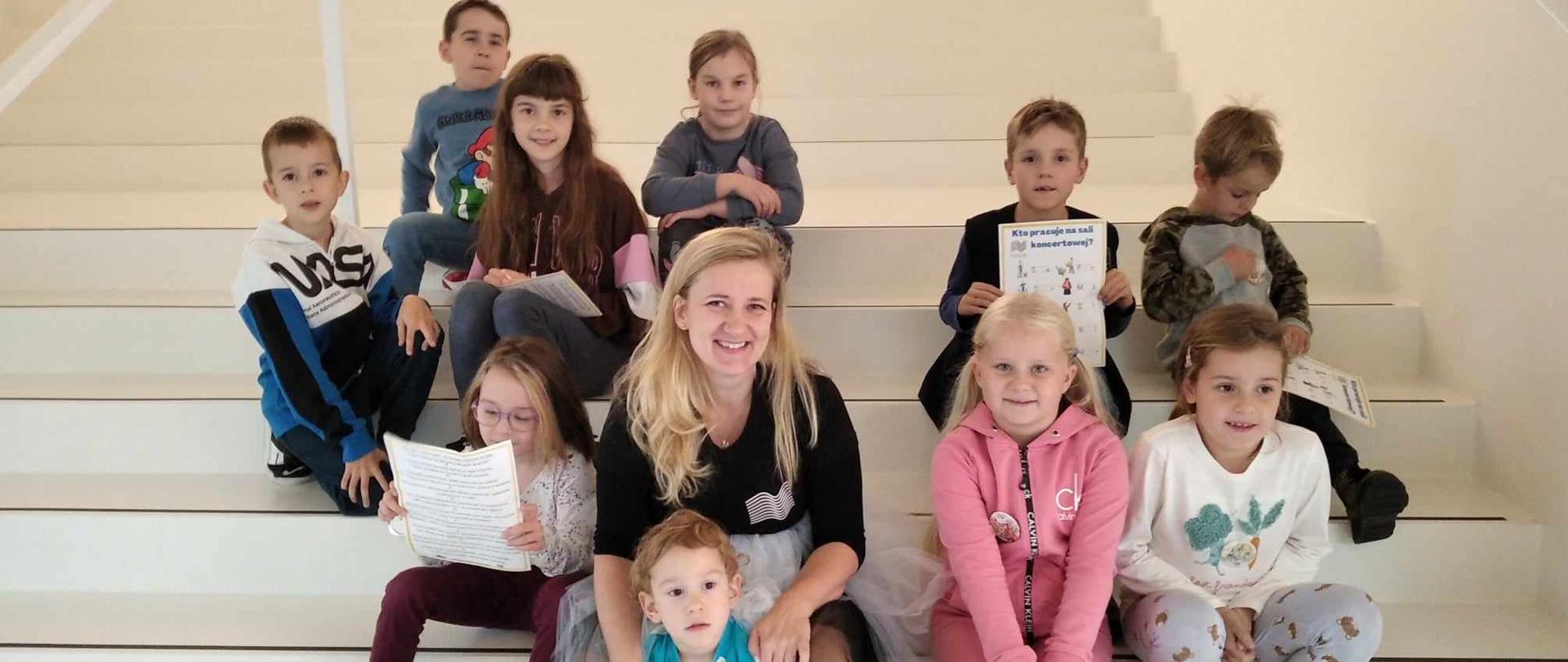Dziesięcioro dzieci siedzi na białych schodach, po środku siedzi kobieta.