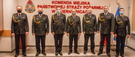 Zdjęcie przedstawia siedmiu strażaków komendy miejskiej w mundurach wyjściowych koloru czarnego na tle czerwonego napisu Komenda Miejska Państwowej Straży Pożarnej w Skierniewicach, loga psp, flag polski i unii europejskiej oraz godła państwowego. 