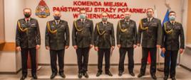 Zdjęcie przedstawia siedmiu strażaków komendy miejskiej w mundurach wyjściowych koloru czarnego na tle czerwonego napisu Komenda Miejska Państwowej Straży Pożarnej w Skierniewicach, loga psp, flag polski i unii europejskiej oraz godła państwowego. 