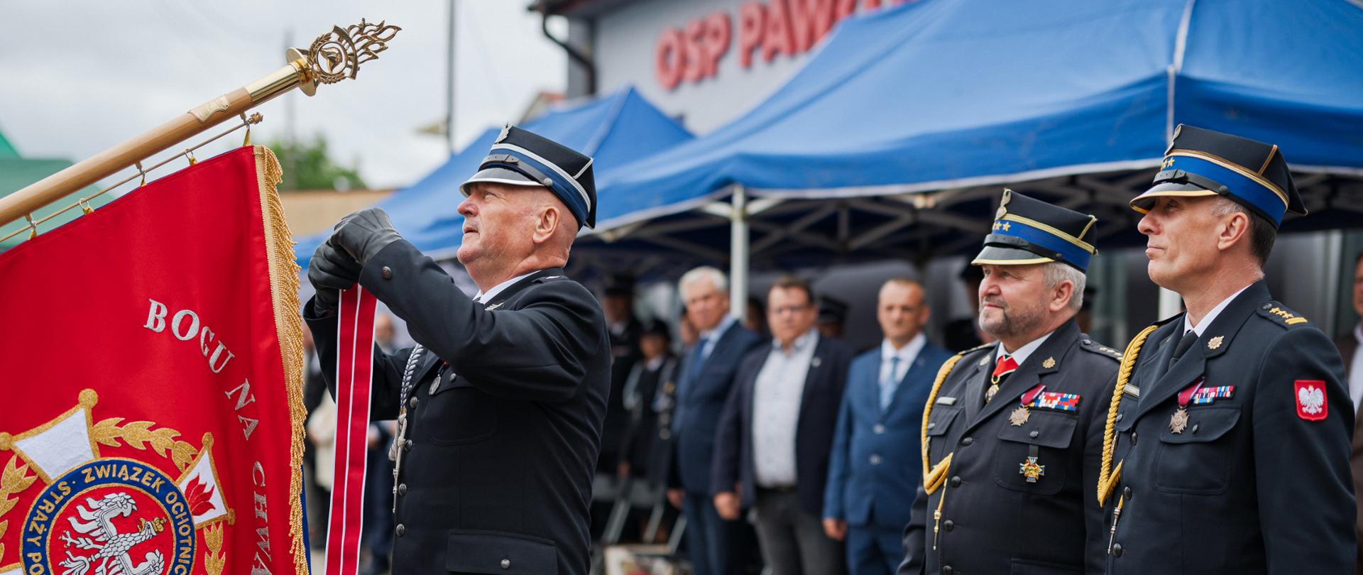 Przedstawiciele straży pożarnej uczestniczą w ceremonii nadania sztandaru podczas obchodów 100-lecia OSP Pawłowo Kościelne. Na pierwszym planie widoczny jest czerwony sztandar jednostki, który odznaczany Złotym Znakiem Związku ZOSP RP. W tle znajdują się zaproszeni goście oraz budynek strażnicy.