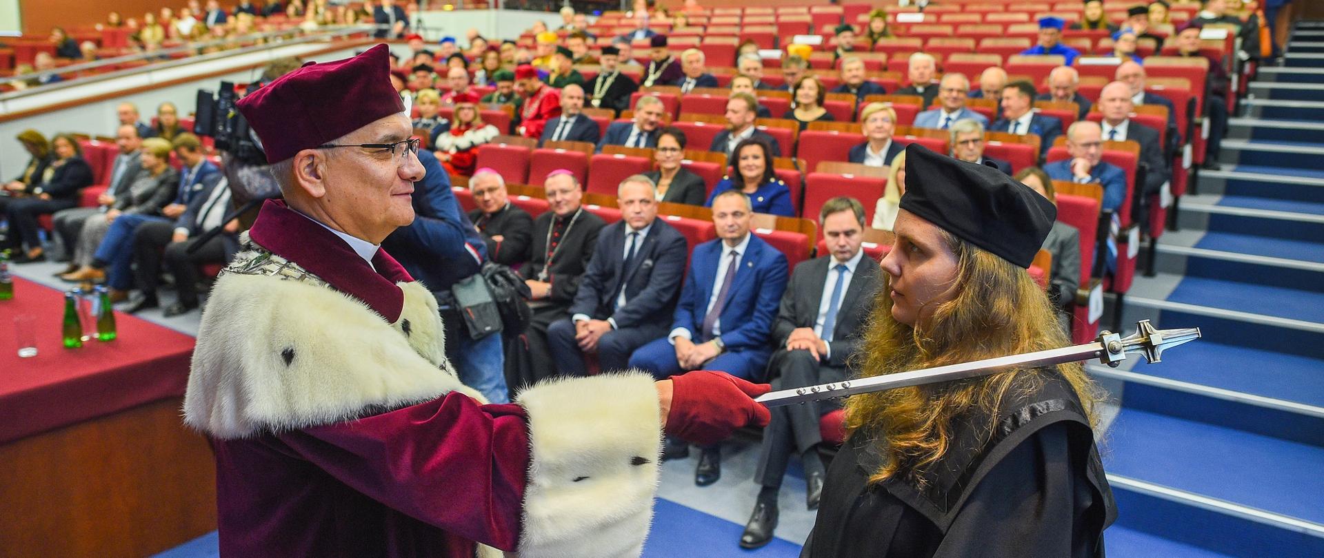 Mężczyzna w czerwonym ceremonialnym stroju dotyka kijkiem ramienia młodej ubranej na czarno dziewczyny w czapce, za nimi sala wypełniona ludźmi.