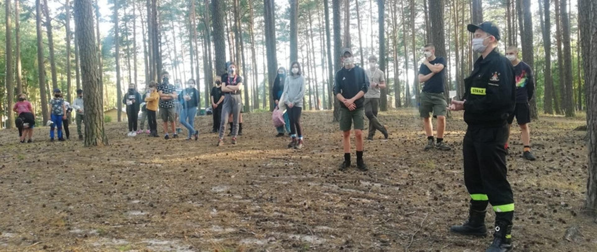 Zdjęcie przedstawia grupę harcerzy w bezpiecznych odstępach, szkolonych przez strażaka. Wszyscy są w maseczkach, plac w lesie