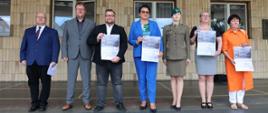 Organizatorzy wydarzenia pozują do zdjęcia pod Opolskim Urzędem Wojewódzkim. Marszałek Szymon Ogłaza, Wojewoda Monika Jurek, Przedstawiciel Instytutu Śląskiego Joanna Siek oraz Prezes Okręgu w Opolu Światowego Związku Żołnierzy Armii Krajowej Krystyna Ejzerman trzymają w rękach plakaty promujące wydarzenie. 