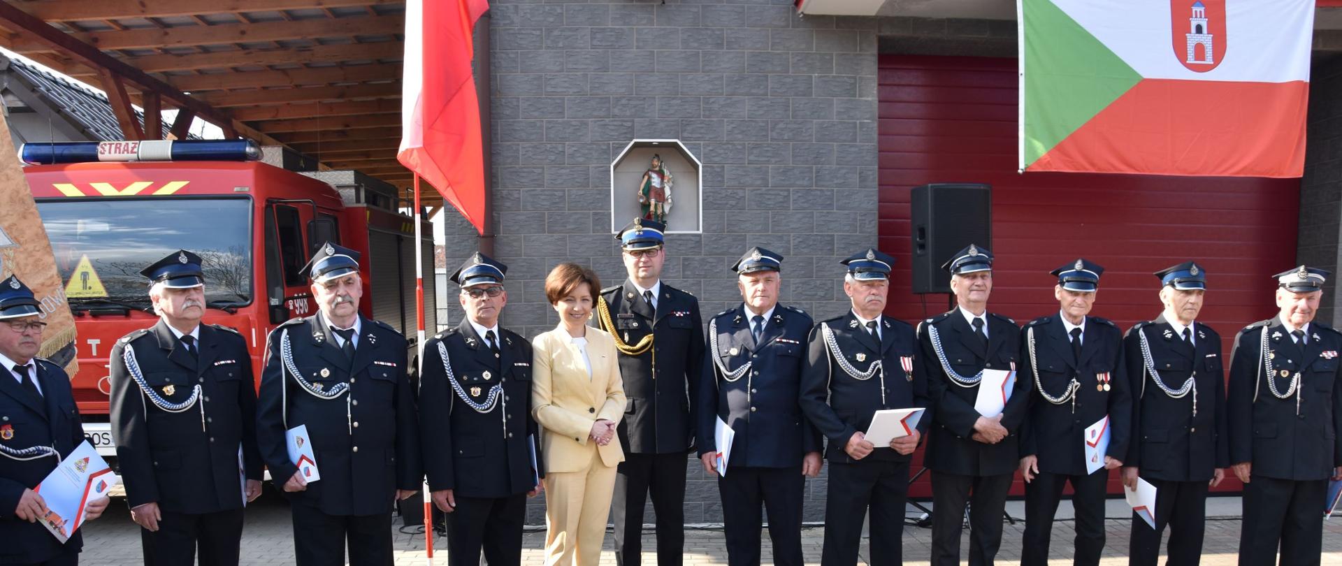 Pani Minister, Komendant Wojewódzki PSP oraz druhowie-emeryci stojący przed gmachem remizy OSP i samochodem strażackim z wręczonymi decyjzami.