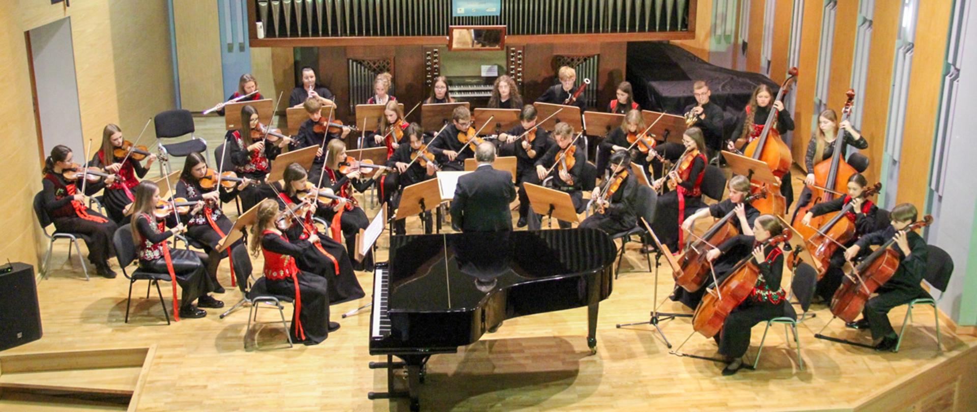 Na zdjęciu widać orkiestrę kameralną grającą w sali koncertowej. Muzycy siedzą w półokręgu wokół dyrygenta, który stoi na środku, tyłem do aparatu. Przed nim stoi fortepian z otwartą pokrywą. Orkiestra składa się głównie z młodych ludzi – widać skrzypków, altowiolistów, wiolonczelistów oraz kontrabasistów. W tle za muzykami znajduje się duży instrument organowy z licznymi metalowymi piszczałkami. Muzycy są elegancko ubrani — część z nich w czarne stroje z czerwonymi i kolorowymi akcentami ludowymi. Sala koncertowa ma drewnianą podłogę i ściany wykończone w jasnych tonach drewna. Atmosfera na zdjęciu jest skupiona i uroczysta — wygląda na to, że orkiestra wykonuje koncert klasyczny.