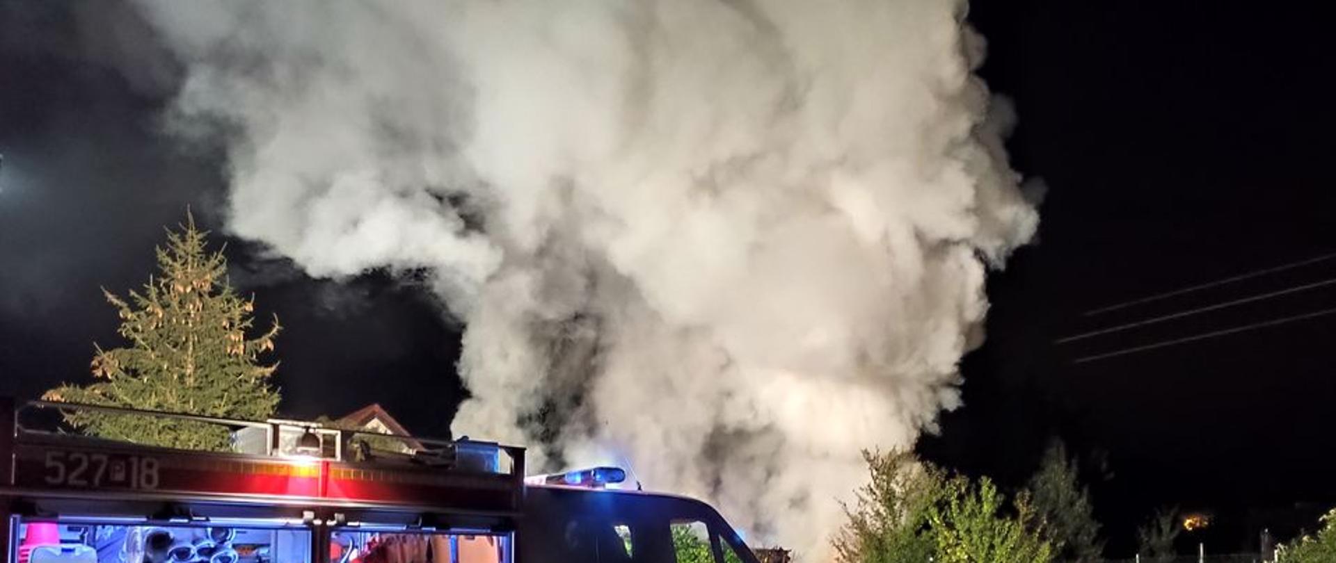 pojazd pożarniczy z otwartymi galotami oświetla teren akcji w tle unoszące się kłęby dymu i pary