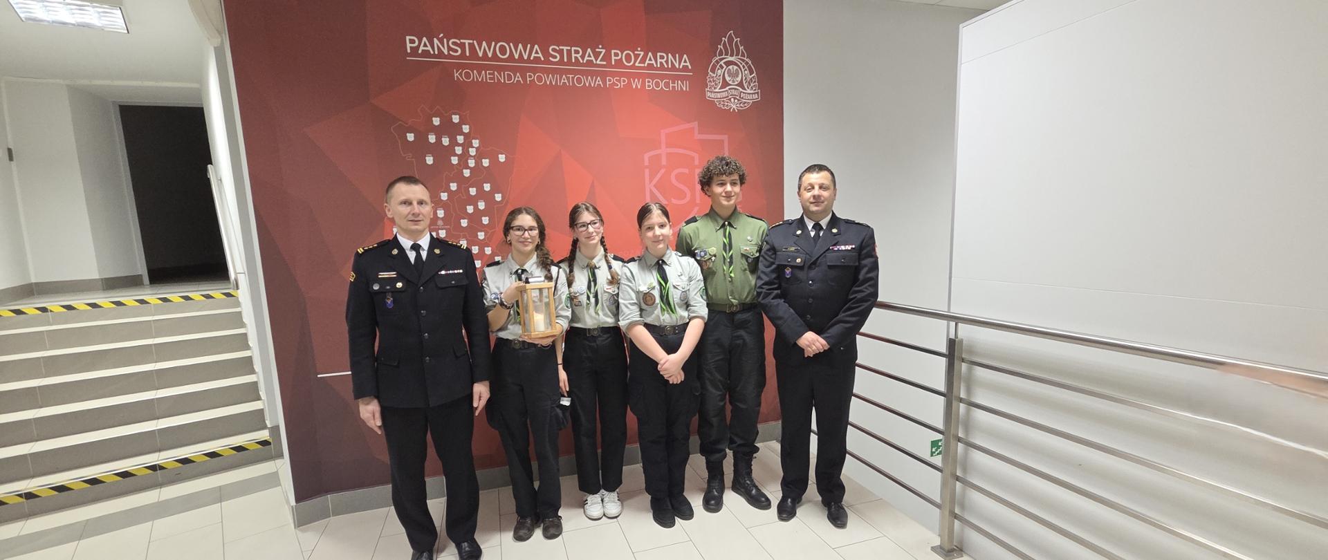 Dwóch strażaków w mundurach galowych wraz z 4 harcerzami pozują do zdjęć w budynku. Za plecami ściana z napisem Komenda Powiatowa PSP w Bochni. Jeden z Harcerzy trzyma w ręce świecznik z zapaloną świecą. 