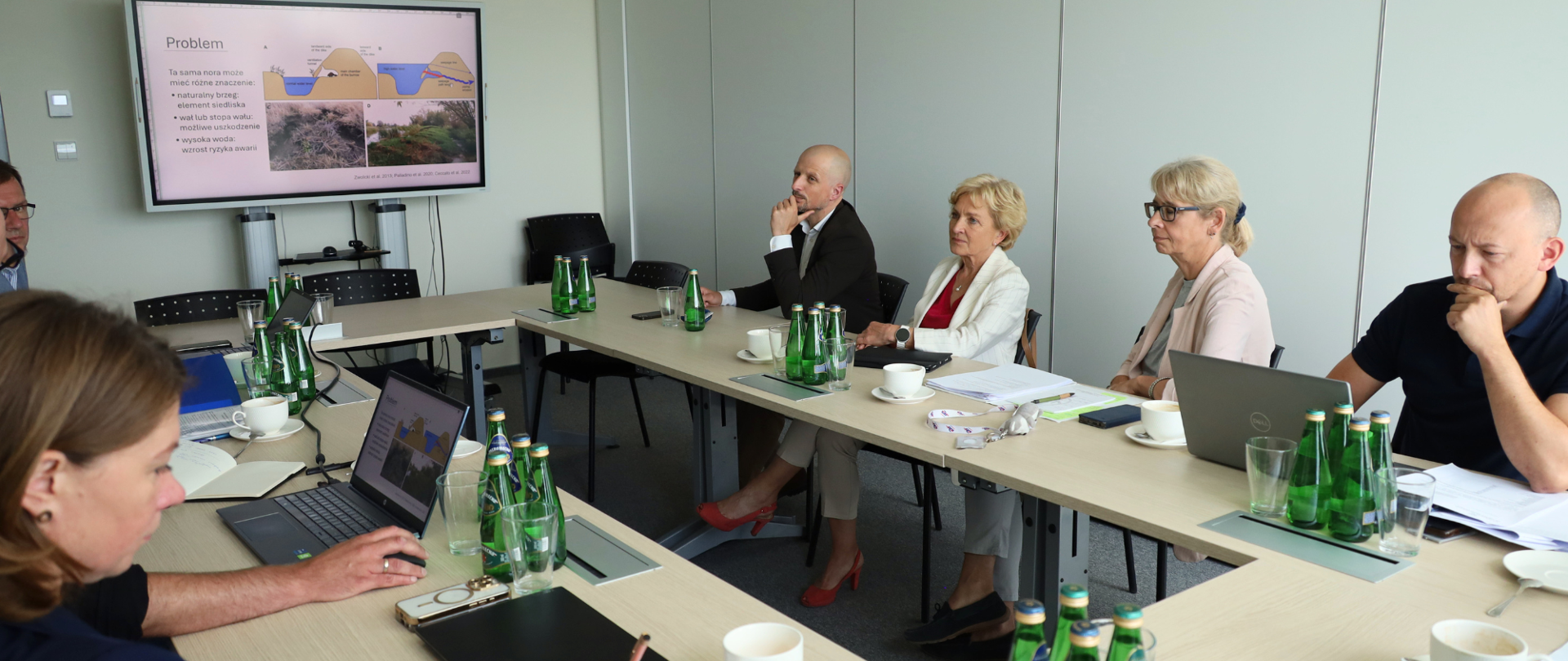 Na zdjęciu przedstawiono spotkanie odbywające się w sali konferencyjnej. Kilka osób siedzi przy ustawionych w kształt litery O stołach, na których znajdują się laptopy, dokumenty, notatniki, filiżanki oraz butelki z wodą. Wszyscy uczestnicy są zwróceni w stronę ekranu zawieszonego na ścianie. Na ekranie wyświetlona jest prezentacja zatytułowana „Problem”, zawierająca schematy oraz fotografie związane z omawianym zagadnieniem. Uczestnicy słuchają prezentacji i analizują przedstawione materiały, a część z nich ma przed sobą komputery z otwartymi dokumentami. Pomieszczenie jest nowoczesne i dobrze oświetlone światłem dziennym. Jasne ściany, uporządkowany układ stołów oraz wyposażenie konferencyjne podkreślają formalny charakter spotkania. Całość sprawia wrażenie merytorycznej dyskusji lub narady dotyczącej projektu.