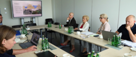 Na zdjęciu przedstawiono spotkanie odbywające się w sali konferencyjnej. Kilka osób siedzi przy ustawionych w kształt litery O stołach, na których znajdują się laptopy, dokumenty, notatniki, filiżanki oraz butelki z wodą. Wszyscy uczestnicy są zwróceni w stronę ekranu zawieszonego na ścianie. Na ekranie wyświetlona jest prezentacja zatytułowana „Problem”, zawierająca schematy oraz fotografie związane z omawianym zagadnieniem. Uczestnicy słuchają prezentacji i analizują przedstawione materiały, a część z nich ma przed sobą komputery z otwartymi dokumentami. Pomieszczenie jest nowoczesne i dobrze oświetlone światłem dziennym. Jasne ściany, uporządkowany układ stołów oraz wyposażenie konferencyjne podkreślają formalny charakter spotkania. Całość sprawia wrażenie merytorycznej dyskusji lub narady dotyczącej projektu.