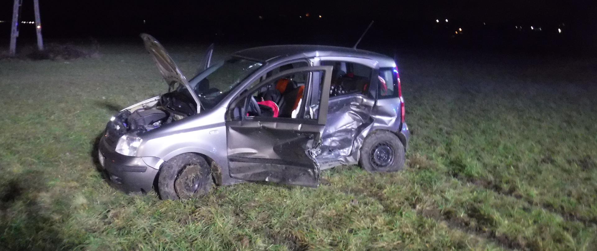 Zdjęcie przedstawia rozbity samochód osobowy marki fiat panda. Samochód ma wgniecione lewe przednie i tylne drzwi. Lewe drzwi są otwarte. Pojazd stoi na podłożu trawiastym. Pojazd ma otwartą klapę przednią. Za pojazdem po lewej stronie widać słup sieci energetycznej.