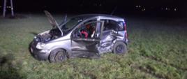 Zdjęcie przedstawia rozbity samochód osobowy marki fiat panda. Samochód ma wgniecione lewe przednie i tylne drzwi. Lewe drzwi są otwarte. Pojazd stoi na podłożu trawiastym. Pojazd ma otwartą klapę przednią. Za pojazdem po lewej stronie widać słup sieci energetycznej.
