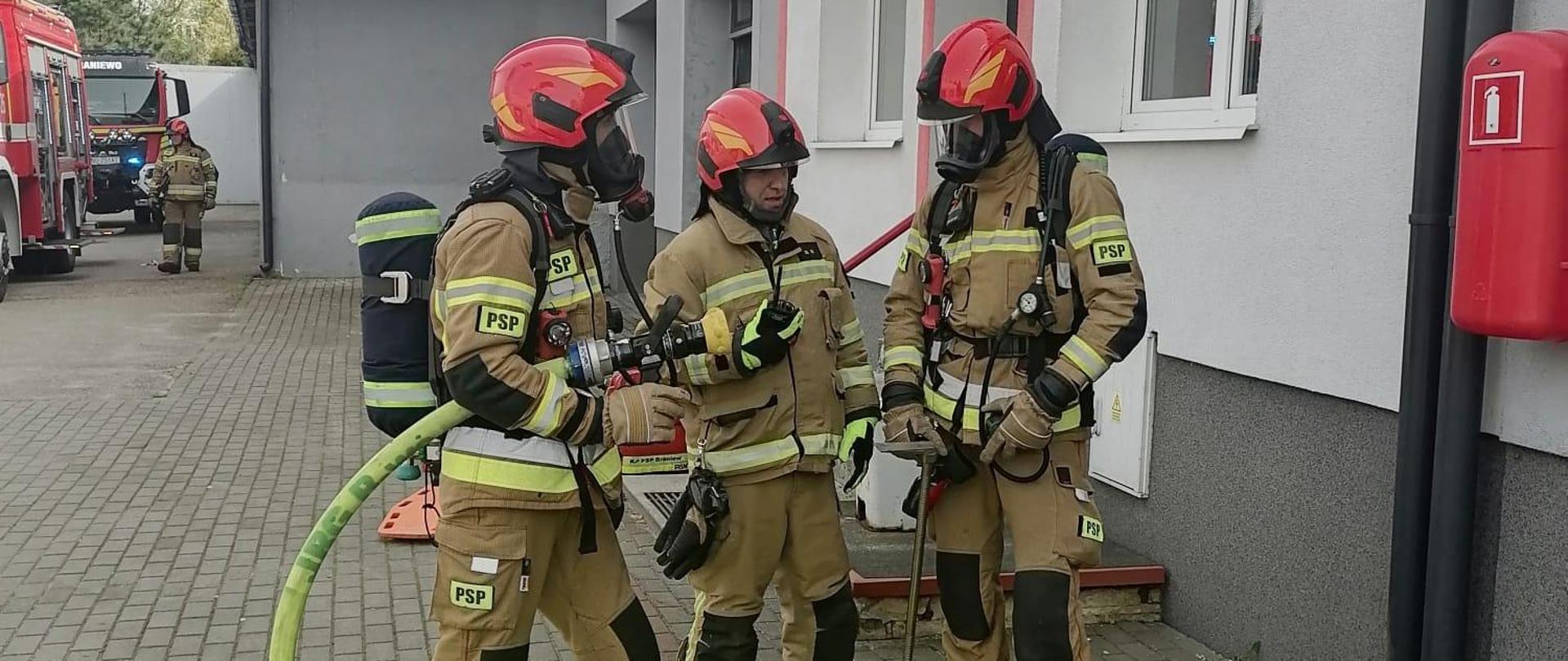 Trzech strażaków przed budynkiem ze sprzętem i wężami. Trwają ćwiczenia.