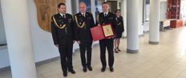 Grupa 4 strażaków w mundurach galowych znajduje się na dużej sali. Jedna osoba trzyma w ręce otwartą teczkę w której znajduje się podziękowanie dla st. kpt, Łukasza Kleckiego za wspólną służbę w PSP.