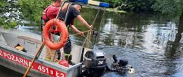 Fotografia przedstawia dwóch ratowników stojących w łodzi motorowej aluminiowej płaskodennej i asekurujących linką ratowniczą barwy żółtej płetwonurka zanurzonego w rzece. Płetwonurek ma na plecach butle powietrzne, kaptur na głowie i maskę do nurkowania. Łódka posiada na części rufowej zamontowany na stalowym stelażu zestaw świateł alarmowych barwy niebieskiej, po lewej stronie stelażu zamontowane jest koło ratunkowe barwy pomarańczowej, o koło i dno rzeki oparty jest bosak. Dookoła rzeki rosną drzewa i krzaki, przy brzegu roślinność i trawa. Niebo pochmurne.