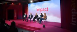Zdjęcie z boku pod kątem, na oświetlonym podwyższeniu z przodu zaciemnionej sali na krzesłach siedzi pięć osób, za nimi na wielkim ekranie napis Impact.