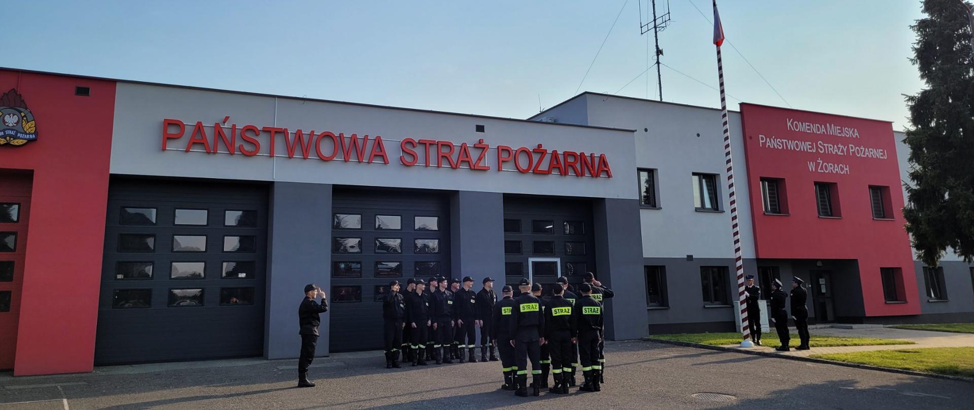 Strażacy stoją na uroczystej zbiórce i salutują podczas podnoszenia flagi państwowej na maszt. W tle budynek Komendy z napisem Państwowa Staż Pożarna oraz obok napis Komenda Miejska Państwowej Straży Pożarnej w Żorach.