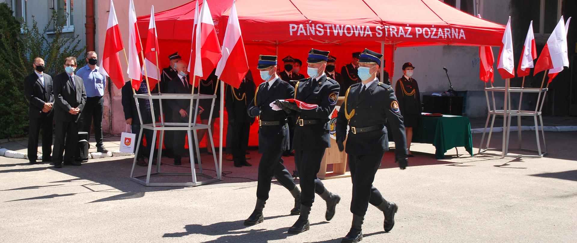 Na pierwszym planie widać trzech strażaków tworzących poczet flagowy, który niesie złożoną flagę, w tle widać czerwony namiot z napisem Państwowa Straż Pożarna w Elblągu, pod którym stoją zaproszeni goście. Wszyscy mają na sobie maseczki i czarne rękawiczki.