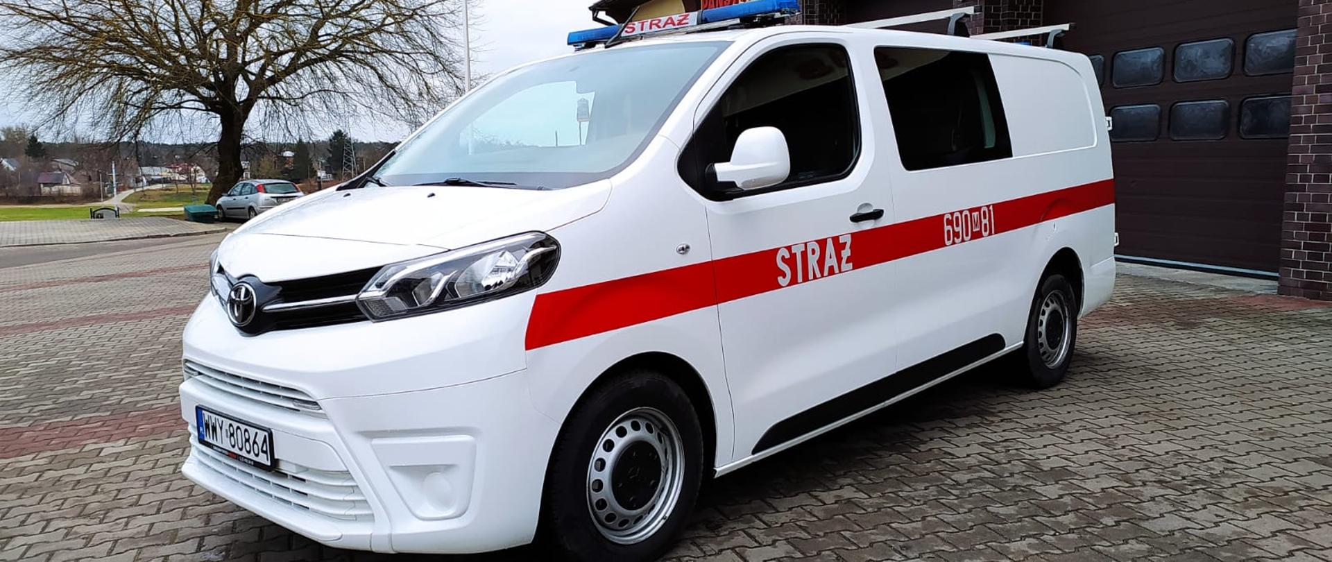 Przed budynkiem Komendy Powiatowej Państwowej Straży Pożarnej w Wyszkowie pojazd Toyota biały z czerwonym pasem z napisem STRAŻ oraz numerami operacyjnymi