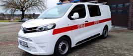 Przed budynkiem Komendy Powiatowej Państwowej Straży Pożarnej w Wyszkowie pojazd Toyota biały z czerwonym pasem z napisem STRAŻ oraz numerami operacyjnymi
