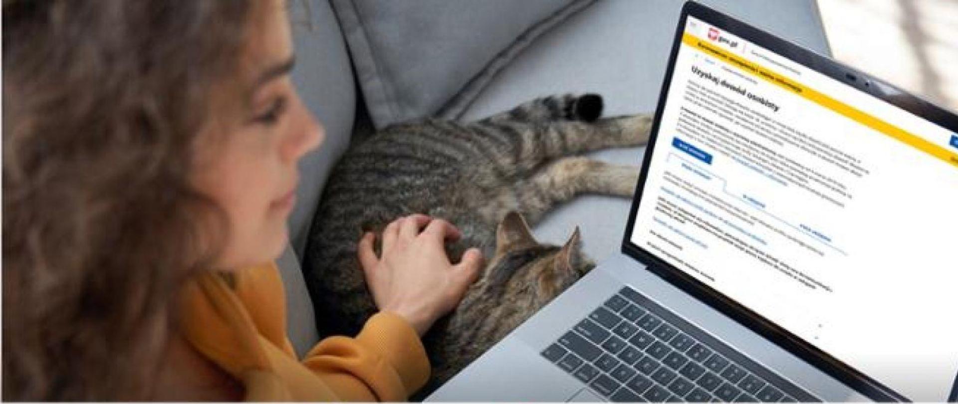 kobieta siedzi przed monitorem komputera, na którym widnieje napis uzyskaj dowód osobisty, obok na kanapie lezy kot