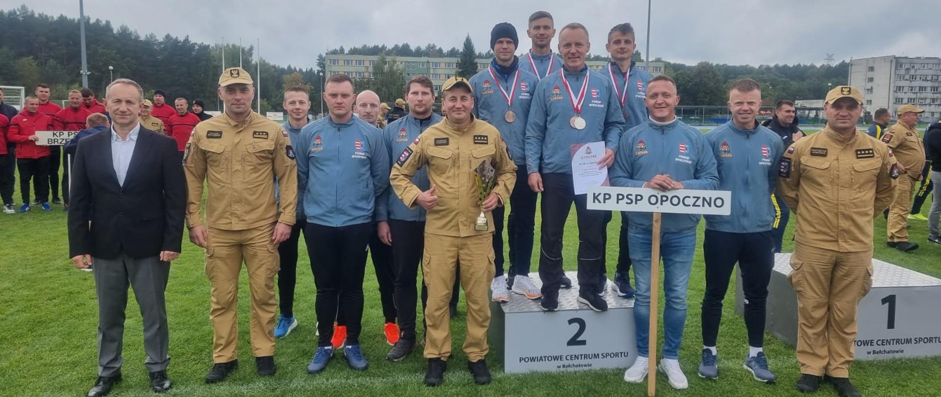 Na zdjęciu trzynastu funkcjonariuszy i jedna osoba cywilna, trzech z nich w umundurowaniu służbowym, czterech z drużyny ma zawieszone medale, dwóch funkcjonariuszy trzyma puchar i dyplom. Drużyna KP PSP Opoczno w dresach, jeden z nich trzyma tablicę z napisem KP PSP Opoczno. W tle inni funkcjonariusze i boisko sportowe.
