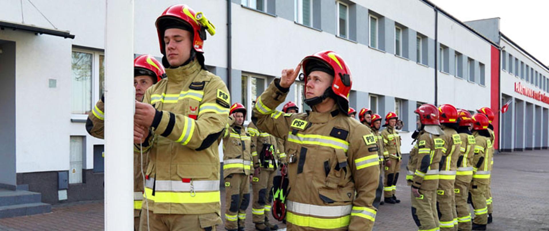 zmiana służbowa strażaków ubranych w mundury koszarowe typu moratex z hełmem na głowie barwy czerwonej stoją na placu przed wejściem do Komendy Powiatowej Staży Pożarnej w Łowiczu, na pierwszym planie trzech strażaków stoi przy maszcie flagowym a jeden strażak wciąga flagę państwową na maszt, po lewej stronie widoczny budynek komendy, plac wyłożony jest kostką brukową, po prawej stronie widoczny namalowany na kostce znak określający miejsce parkingowe dla osób niepełnosprawnych w postaci dużego niebieskiego kwadratu z białą obwódką i symbolem osoby siedzącej na wózku inwalidzkim