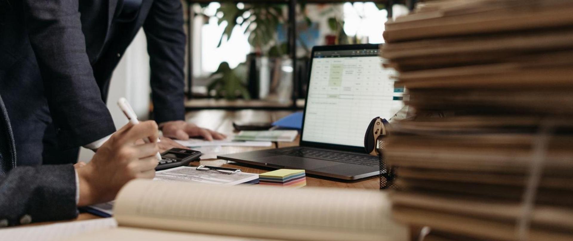 Dwie osoby opierają swoje dłonie na biurku z dokumentami i laptopem